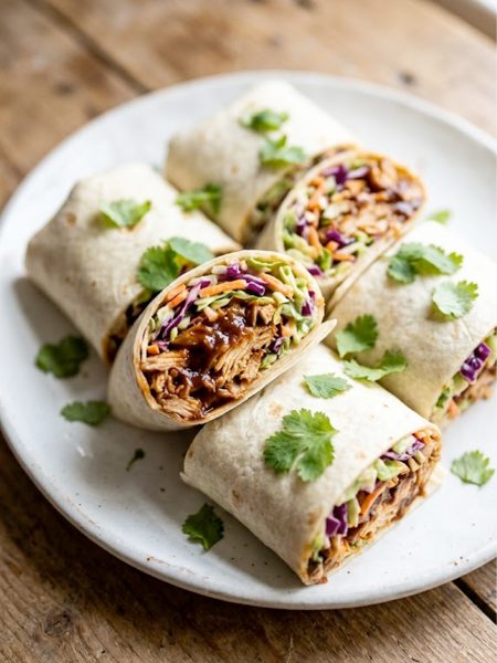 Four BBQ chicken coleslaw wraps cut in half and arranged on a white plate, showing smoky shredded chicken and creamy coleslaw filling, garnished with fresh cilantro