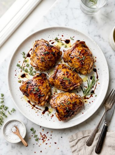 Juicy balsamic glazed chicken thighs on a white ceramic plate topped with fresh thyme and rosemary sprigs on a marble surface