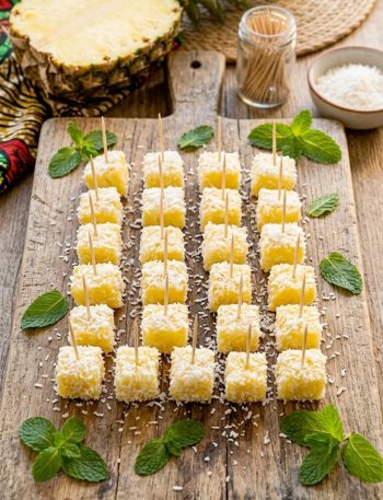 Boozy pina colada bites coated in coconut flakes served on a rustic wooden board with fresh pineapple and mint garnish