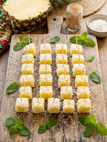 Boozy pina colada bites coated in coconut flakes served on a rustic wooden board with fresh pineapple and mint garnish