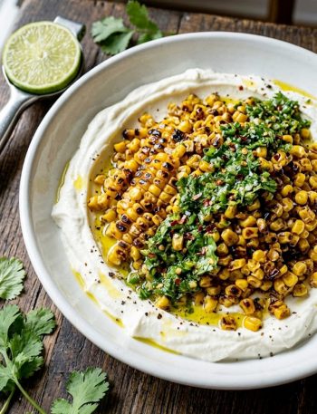 A wide shallow white bowl filled with deeply charred golden corn kernels piled over a smooth layer of whipped feta cheese, topped with a vibrant chili lime cilantro dressing and red chili flakes
