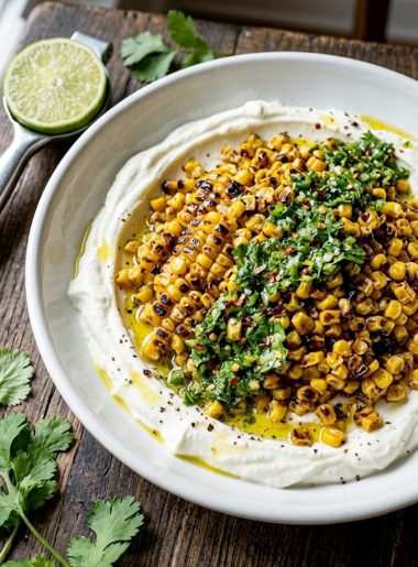 A wide shallow white bowl filled with deeply charred golden corn kernels piled over a smooth layer of whipped feta cheese, topped with a vibrant chili lime cilantro dressing and red chili flakes