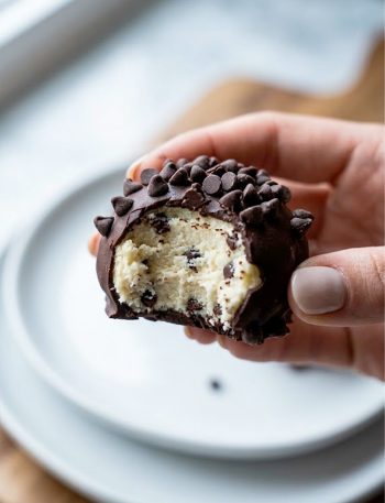 A hand holding a single cheesecake keto fat bomb coated in dark chocolate and covered with sugar-free mini chocolate chips showing the creamy cheesecake interior where it has been bitten into