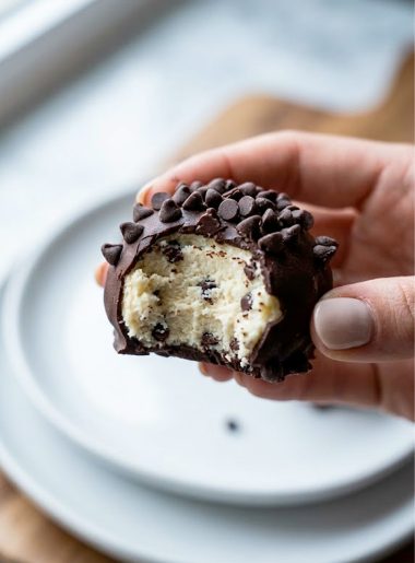 A hand holding a single cheesecake keto fat bomb coated in dark chocolate and covered with sugar-free mini chocolate chips showing the creamy cheesecake interior where it has been bitten into