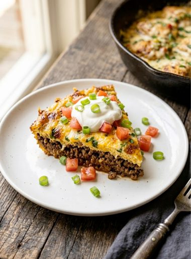 A slice of cheesy keto taco casserole on a white plate topped with sour cream, diced fresh tomatoes and sliced green onions showing layers of seasoned beef and golden egg and cheese base