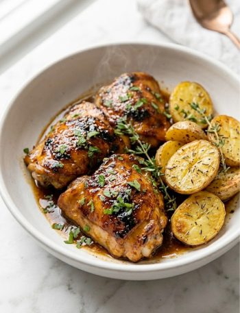 Classic honey BBQ chicken thighs with a sticky caramelized glaze served alongside golden roasted baby potatoes garnished with fresh herbs in a wide white bowl on a wooden table