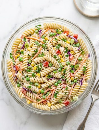 A large glass bowl filled with creamy crack pasta salad made with rotini pasta shredded carrots sweet peas corn red bell pepper and red cabbage tossed in a tangy ranch inspired dressing