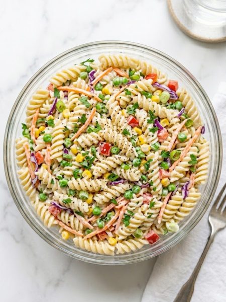 A large glass bowl filled with creamy crack pasta salad made with rotini pasta shredded carrots sweet peas corn red bell pepper and red cabbage tossed in a tangy ranch inspired dressing