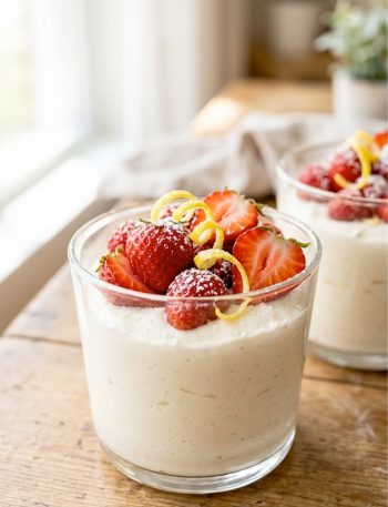 Creamy keto lemon cheesecake mousse served in a clear glass topped with fresh strawberries and lemon zest curls on a rustic wooden table