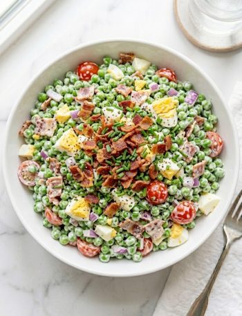 A large white ceramic bowl of creamy pea salad with bacon loaded with sweet green peas, crumbled crispy bacon, chopped hard-boiled eggs, halved cherry tomatoes, and fresh chives tossed in a rich tangy dressing on a white marble surface