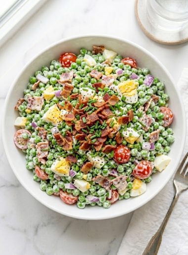 A large white ceramic bowl of creamy pea salad with bacon loaded with sweet green peas, crumbled crispy bacon, chopped hard-boiled eggs, halved cherry tomatoes, and fresh chives tossed in a rich tangy dressing on a white marble surface