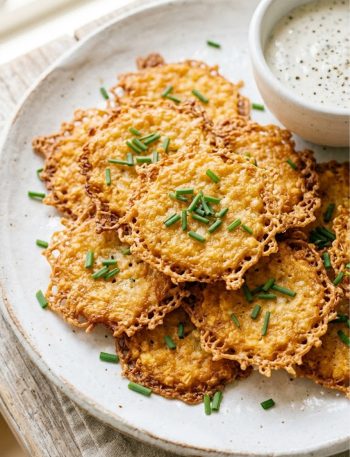 Golden crispy baked cottage cheese chips arranged on a white plate garnished with fresh chives, with a small bowl of creamy white dipping sauce in the background, on a rustic wooden surface