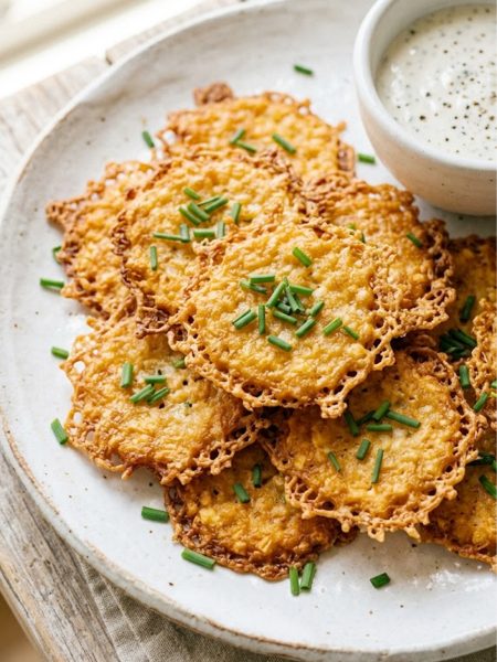 Golden crispy baked cottage cheese chips arranged on a white plate garnished with fresh chives, with a small bowl of creamy white dipping sauce in the background, on a rustic wooden surface