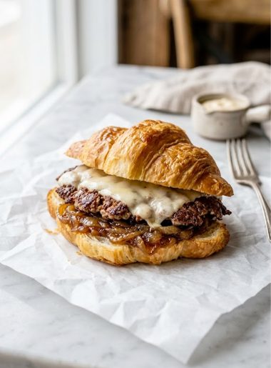 A golden toasted croissant smash burger with melted cheese and caramelized onions on parchment paper