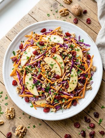 A large white plate filled with crunchy apple carrot salad topped with toasted walnuts, dried cranberries, fresh parsley, and creamy citrus dressing on a wooden table