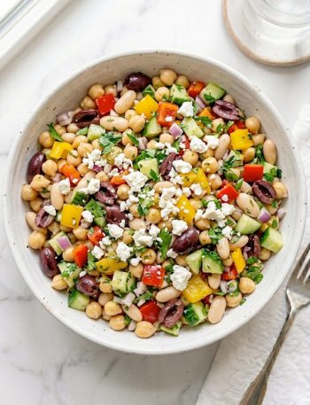 A large white ceramic bowl of dense bean salad loaded with chickpeas, white cannellini beans, black olives, diced red and yellow bell peppers, cucumber, red onion, fresh parsley, and crumbled feta cheese tossed in a bright tangy vinaigrette on a white marble surface
