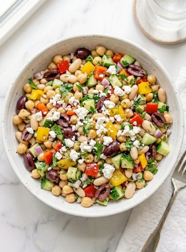 A large white ceramic bowl of dense bean salad loaded with chickpeas, white cannellini beans, black olives, diced red and yellow bell peppers, cucumber, red onion, fresh parsley, and crumbled feta cheese tossed in a bright tangy vinaigrette on a white marble surface