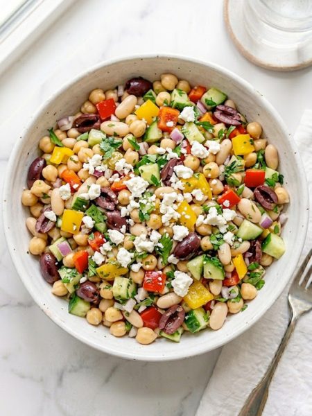 A large white ceramic bowl of dense bean salad loaded with chickpeas, white cannellini beans, black olives, diced red and yellow bell peppers, cucumber, red onion, fresh parsley, and crumbled feta cheese tossed in a bright tangy vinaigrette on a white marble surface