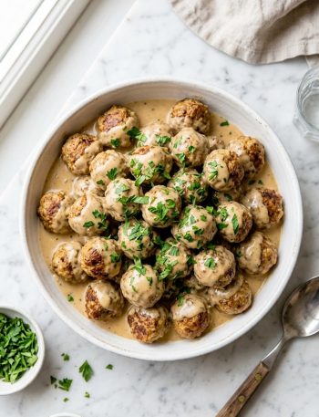 Golden baked Easy Swedish meatballs smothered in a rich creamy sauce garnished with fresh parsley in a white ceramic serving dish