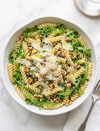 A large white bowl filled with lemon arugula pasta salad made with fusilli pasta fresh peppery arugula capers toasted walnuts and topped with generous shaved parmesan and cracked black pepper