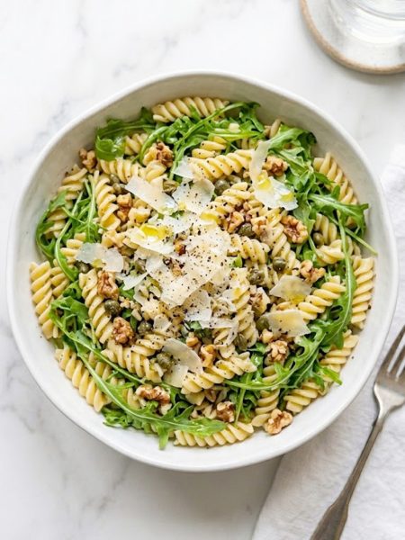 A large white bowl filled with lemon arugula pasta salad made with fusilli pasta fresh peppery arugula capers toasted walnuts and topped with generous shaved parmesan and cracked black pepper