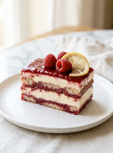 A plated slice of easy raspberry tiramisu showing distinct layers of mascarpone cream and raspberry soaked ladyfingers topped with fresh raspberries and a lemon slice on a white plate