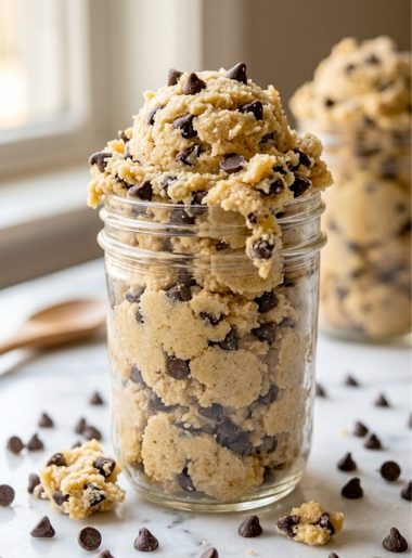 A glass mason jar overflowing with pale golden edible keto cookie dough studded with dark sugar-free chocolate chips on a white marble surface with extra chocolate chips scattered around