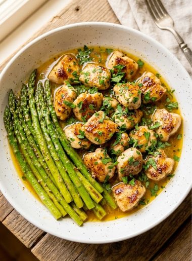 A white ceramic serving bowl filled with golden seared garlic butter chicken bites glistening in a rich golden butter sauce with fresh parsley, served alongside bright green asparagus spears on a rustic wooden surface