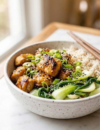 Close up of a white speckled ceramic bowl filled with ginger honey chicken, fluffy basmati rice, and sauteed bok choy garnished with green onions and sesame seeds