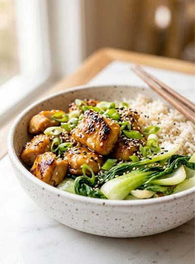 Close up of a white speckled ceramic bowl filled with ginger honey chicken, fluffy basmati rice, and sauteed bok choy garnished with green onions and sesame seeds