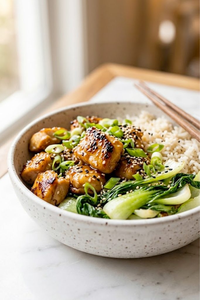 Close up of a white speckled ceramic bowl filled with ginger honey chicken, fluffy basmati rice, and sauteed bok choy garnished with green onions and sesame seeds