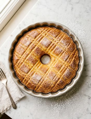 Golden French butter cake with a crosshatch pattern and sugar crust in a round silver baking pan on a marble surface