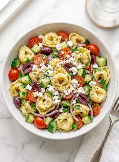 A large white ceramic bowl of Greek tortellini pasta salad filled with cheese tortellini, halved cherry tomatoes, kalamata olives, diced cucumber, thinly sliced red onion, fresh parsley, and generous crumbles of feta cheese tossed in a bright Greek vinaigrette on a white marble surface
