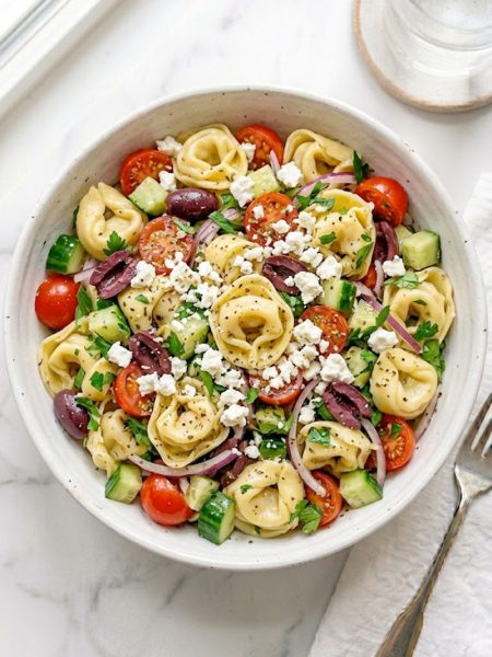 A large white ceramic bowl of Greek tortellini pasta salad filled with cheese tortellini, halved cherry tomatoes, kalamata olives, diced cucumber, thinly sliced red onion, fresh parsley, and generous crumbles of feta cheese tossed in a bright Greek vinaigrette on a white marble surface