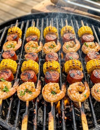 Multiple grilled shrimp and sausage skewers with charred corn rounds cooking on a hot outdoor grill grate seasoned with Cajun spice rub