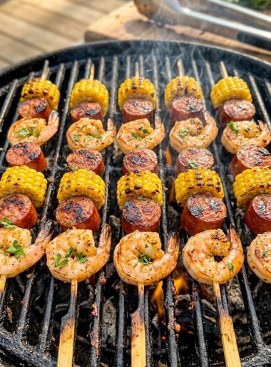 Multiple grilled shrimp and sausage skewers with charred corn rounds cooking on a hot outdoor grill grate seasoned with Cajun spice rub