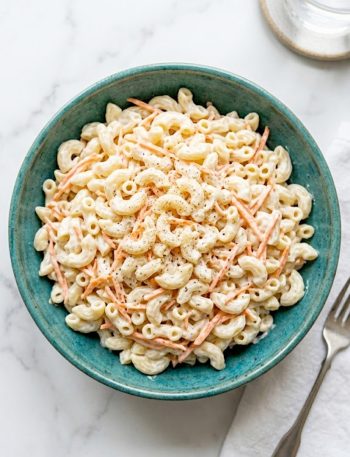 A large teal ceramic bowl filled with creamy Hawaiian macaroni salad made with elbow macaroni, shredded carrots, and a rich mayo dressing, garnished with white pepper on a white marble surface