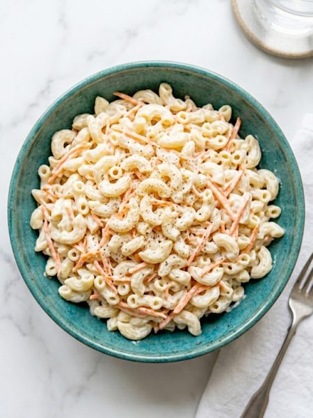 A large teal ceramic bowl filled with creamy Hawaiian macaroni salad made with elbow macaroni, shredded carrots, and a rich mayo dressing, garnished with white pepper on a white marble surface