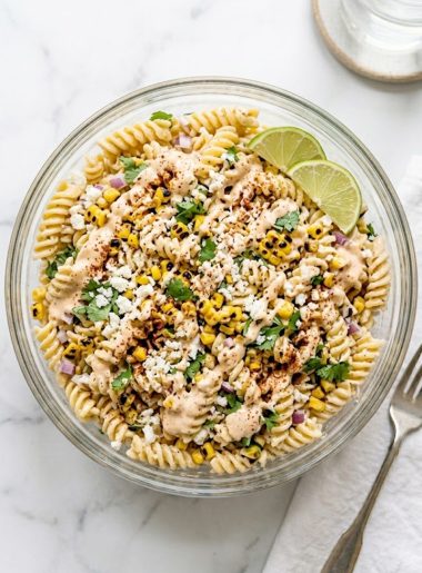 A large glass bowl of high protein elote pasta salad topped with crumbled cotija cheese, fresh cilantro, and chili powder with lime wedges on the side