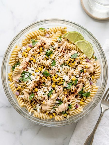 A large glass bowl of high protein elote pasta salad topped with crumbled cotija cheese, fresh cilantro, and chili powder with lime wedges on the side