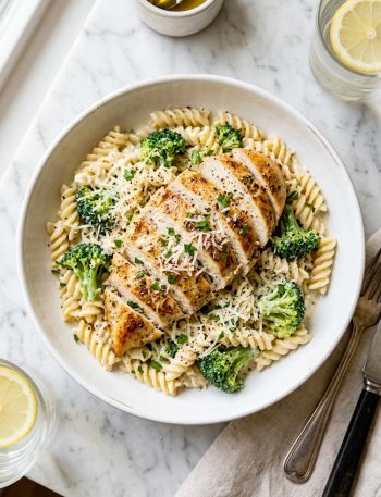 High protein chicken pasta with golden seared sliced chicken breast over creamy garlic parmesan rotini pasta and tender broccoli florets garnished with fresh parsley and grated parmesan in a white ceramic bowl