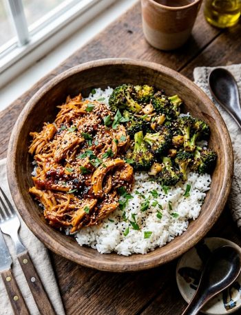 High protein shredded chicken bowl with saucy pulled chicken roasted broccoli florets and fluffy white rice garnished with sesame seeds and fresh herbs in a wooden bowl