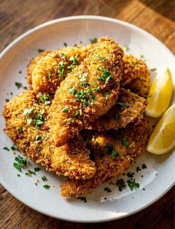 Homemade crispy lemon herb chicken tenders piled high on a white plate with a golden panko crust garnished with fresh parsley and lemon wedges
