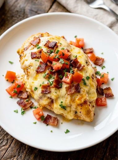 Homemade Monterey chicken on a white plate topped with melted cheese, crispy bacon, diced tomatoes and fresh herbs