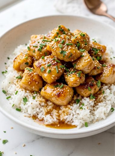 Overhead close up of honey garlic chicken dinner with perfectly glazed golden chicken pieces piled over fluffy white rice in a white ceramic bowl garnished with fresh chives parsley and sesame seeds