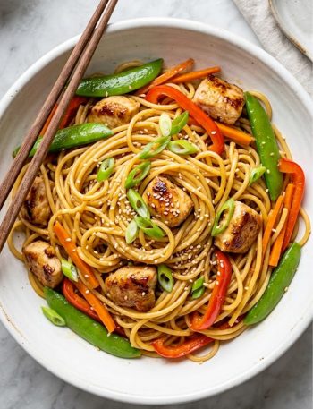 Overhead view of honey garlic chicken noodles with tender chicken, snap peas, red bell pepper, and sesame seeds in a white ceramic bowl