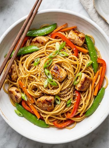 Overhead view of honey garlic chicken noodles with tender chicken, snap peas, red bell pepper, and sesame seeds in a white ceramic bowl