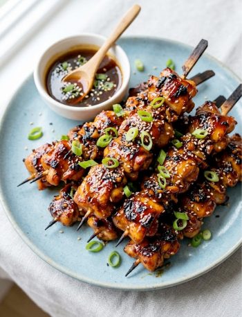 Overhead view of sticky honey garlic chicken skewers piled on a blue ceramic plate garnished with sliced green onions and sesame seeds
