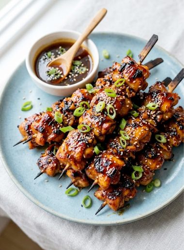 Overhead view of sticky honey garlic chicken skewers piled on a blue ceramic plate garnished with sliced green onions and sesame seeds
