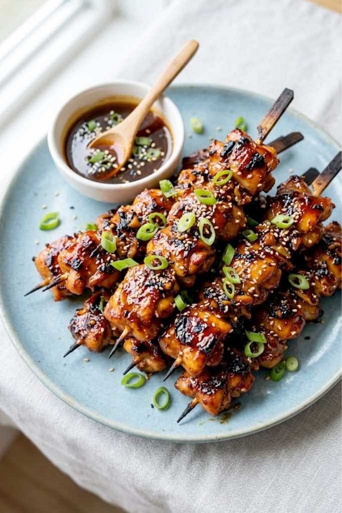 Overhead view of sticky honey garlic chicken skewers piled on a blue ceramic plate garnished with sliced green onions and sesame seeds